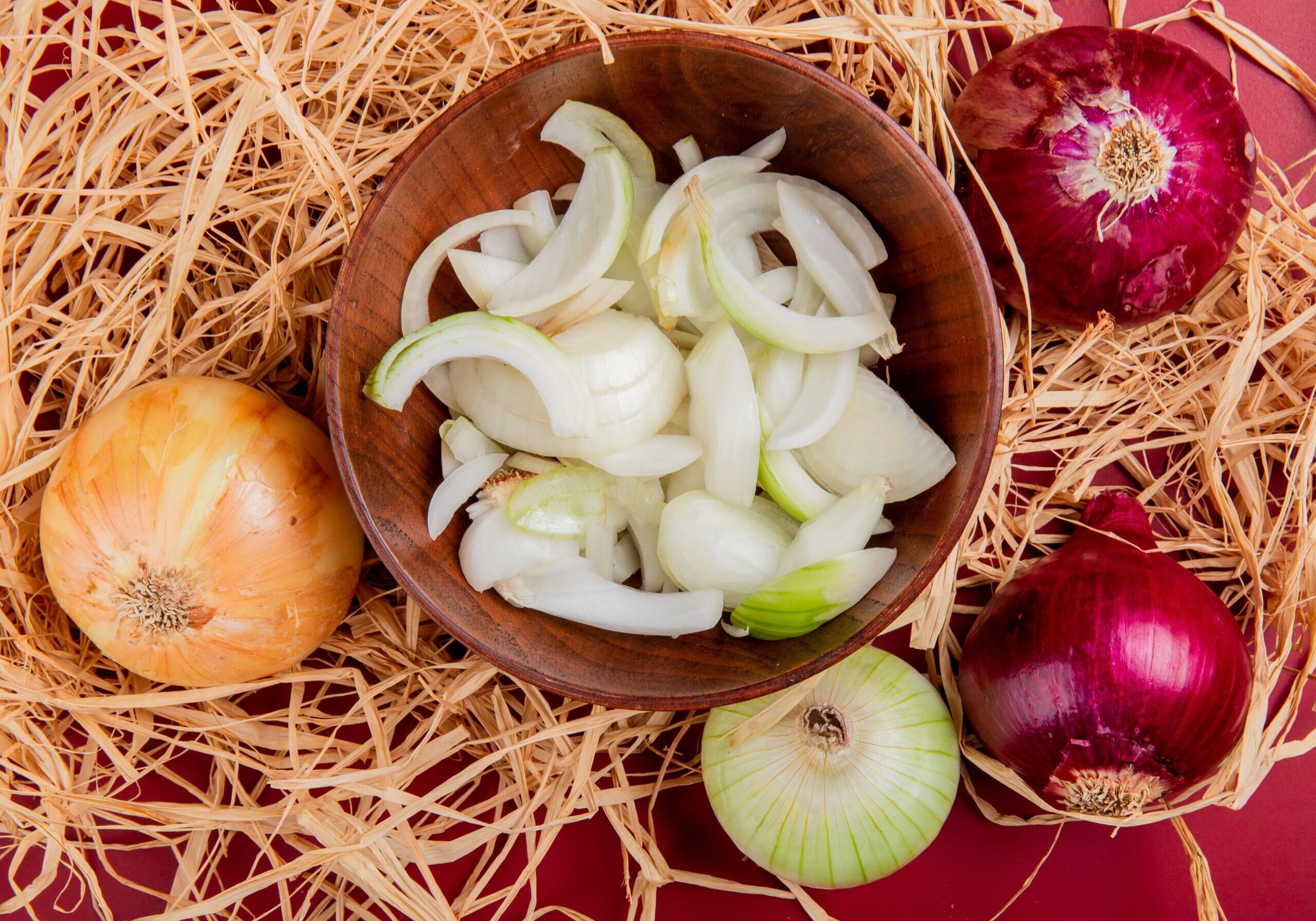 top view of sliced white onion in bowl with sweet, red and whole white one on straw and red background